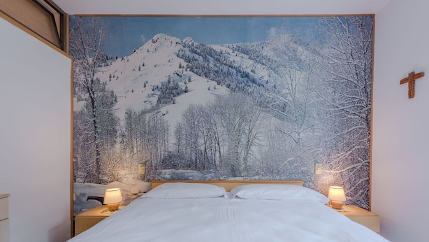 A bedroom with a large bed, two bedside tables with lamps, a wooden headboard, and a wall tapestry of a snowy mountain landscape.