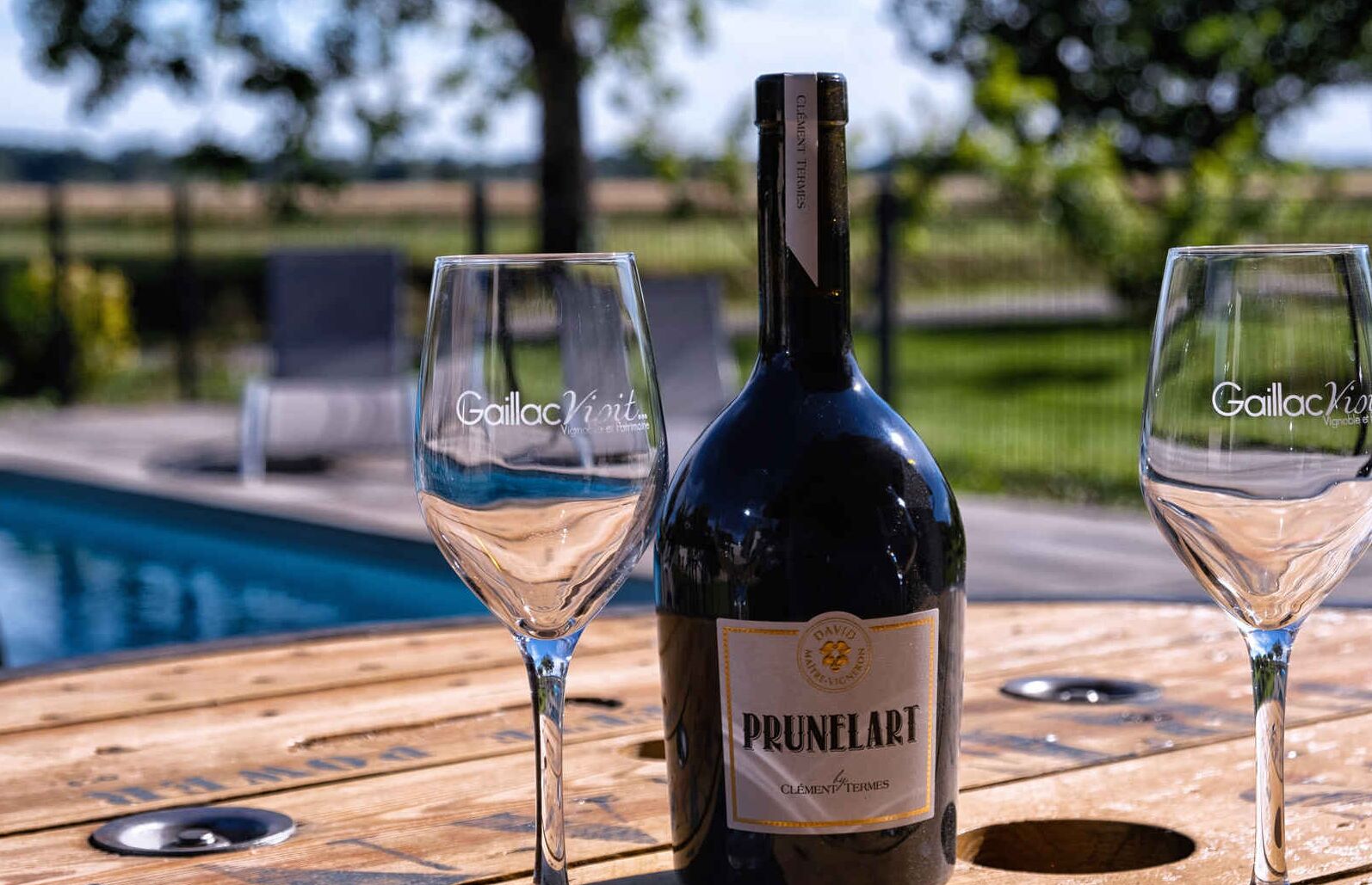 A bottle of Prunelart wine and two glasses on a wooden table by a pool.