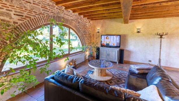 Un salon chaleureux avec une arche en pierre, des poutres en bois au plafond, un canapé en cuir, une table basse et un téléviseur.