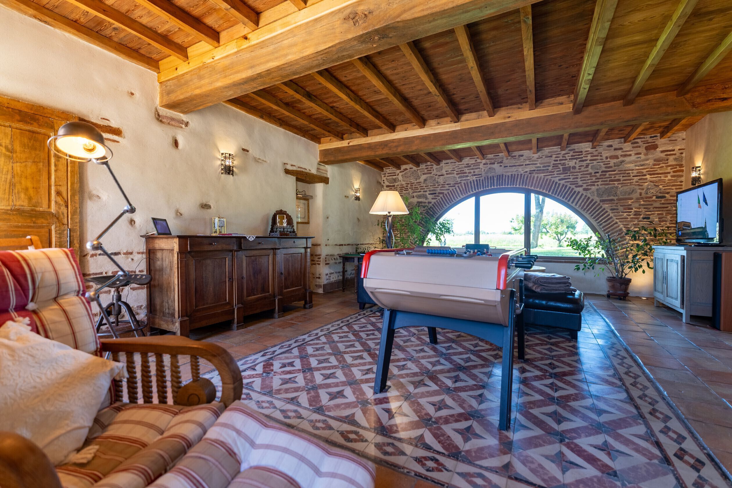 A rustic interior with exposed wooden beams, a stone wall, a large arched window, and a cozy seating area.