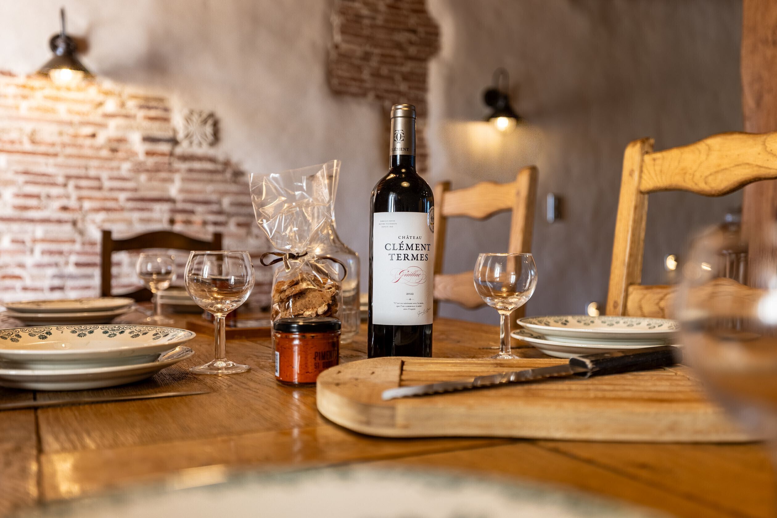 Ein Esstisch mit einer Flasche Château Clément Terme Wein, Weingläsern, Tellern und einem Holzschneidebrett.