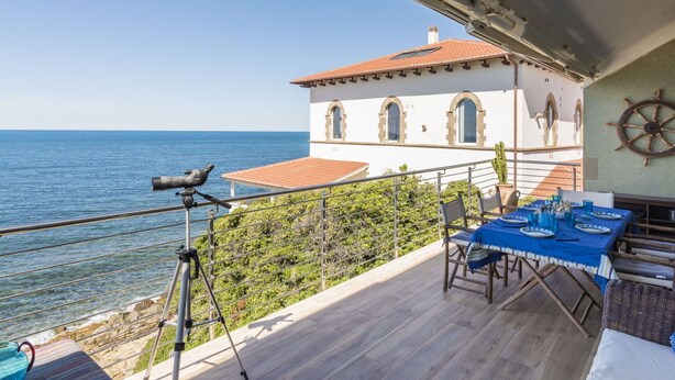 A balcony with a table set for dining, overlooking a coastal view with a building featuring arched windows and a ship's wheel. - Cosy apartment for 7 guests with WIFI, TV, terrace and pets allowed (Civitavecchia)