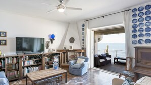 A living room with a TV, bookshelves, a coffee table, and a view of the sea through a large window. - Cosy apartment for 7 guests with WIFI, TV, terrace and pets allowed (Civitavecchia)
