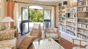 Een kamer met een groot open raam, rieten meubels en een boekenplank vol boeken.