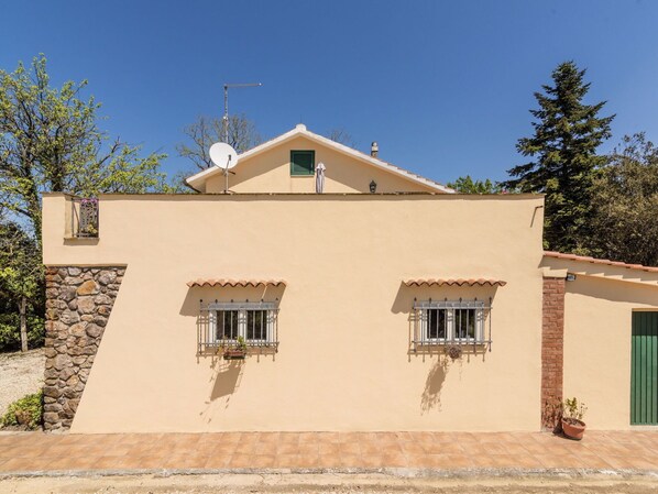 A two-story building with a beige facade, stone wall on the left, and a green door on the right. There are two windows with white shutters and a satellite dish on the roof.