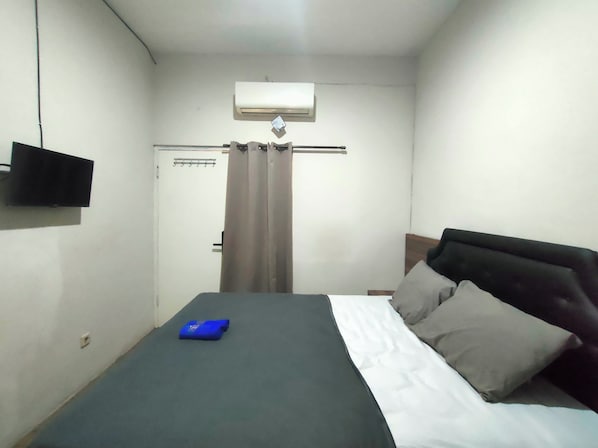 A small, simple room with a bed, a flat-screen TV mounted on the wall, and an air conditioning unit above the door.
