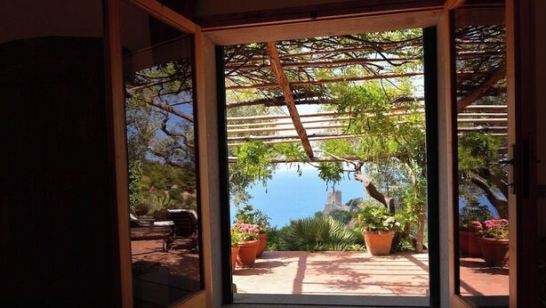 A room with a large sliding glass door leading to an outdoor patio with a pergola, potted plants, and a view of the sea.