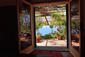 Una habitación con un amplio ventanal corredizo que da a un patio exterior con pérgola, plantas en macetas y vistas al mar.