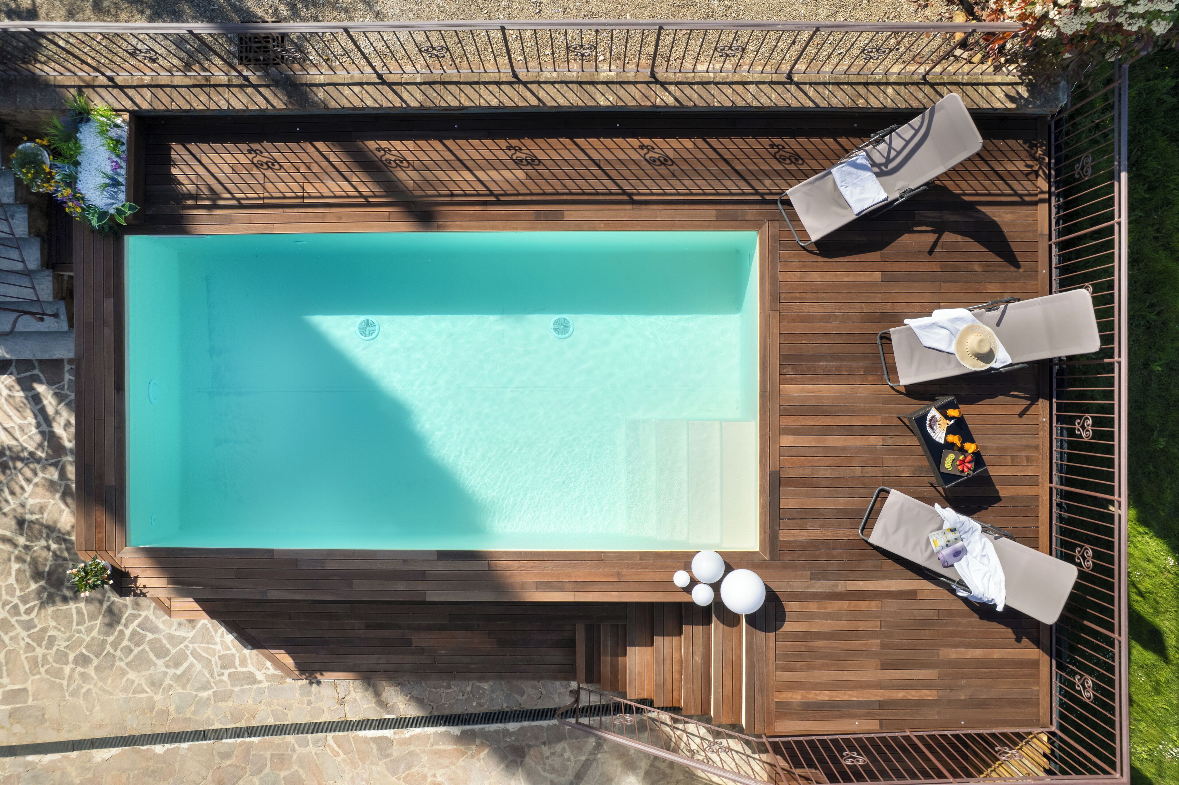 An aerial view of a rectangular swimming pool with a wooden deck surrounding it. There are lounge chairs and a small table with a drink on the deck.
