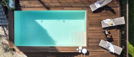 An aerial view of a rectangular swimming pool with a wooden deck surrounding it. There are lounge chairs and a small table with a drink on the deck.