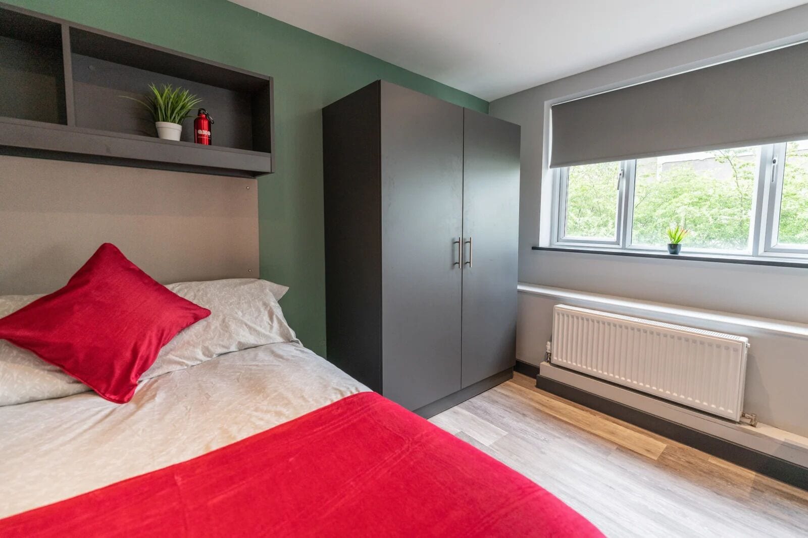 A bedroom with a bed, a red pillow, a grey wardrobe, a window with a view of greenery, and a radiator.