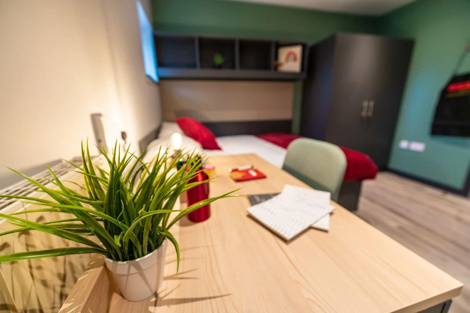 A modern room with a wooden table, a potted plant, a red mug, and a bed with red pillows.
