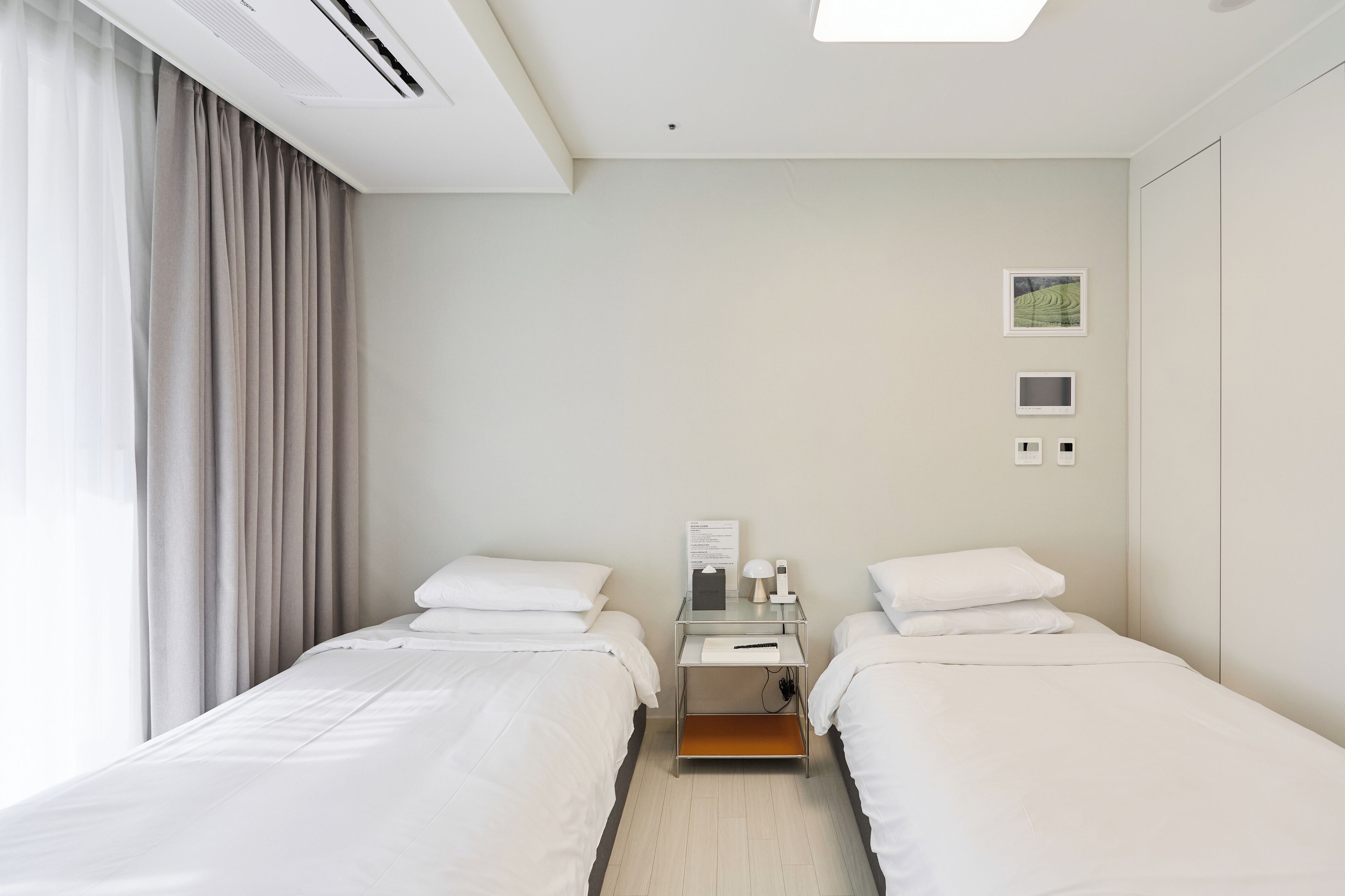 A hotel room with two beds, a bedside table with a lamp, a small desk, and a window with curtains.