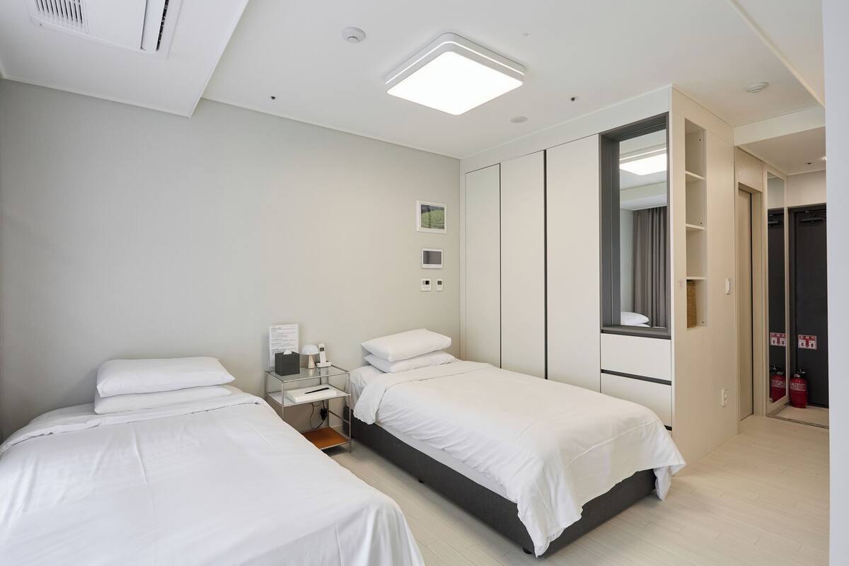 A hotel room with two single beds, a bedside table with a lamp, a small desk with a chair, a wardrobe, and a large mirror.