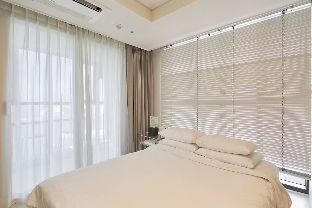 A bedroom with a large bed, a nightstand, a lamp, and a window with blinds.
