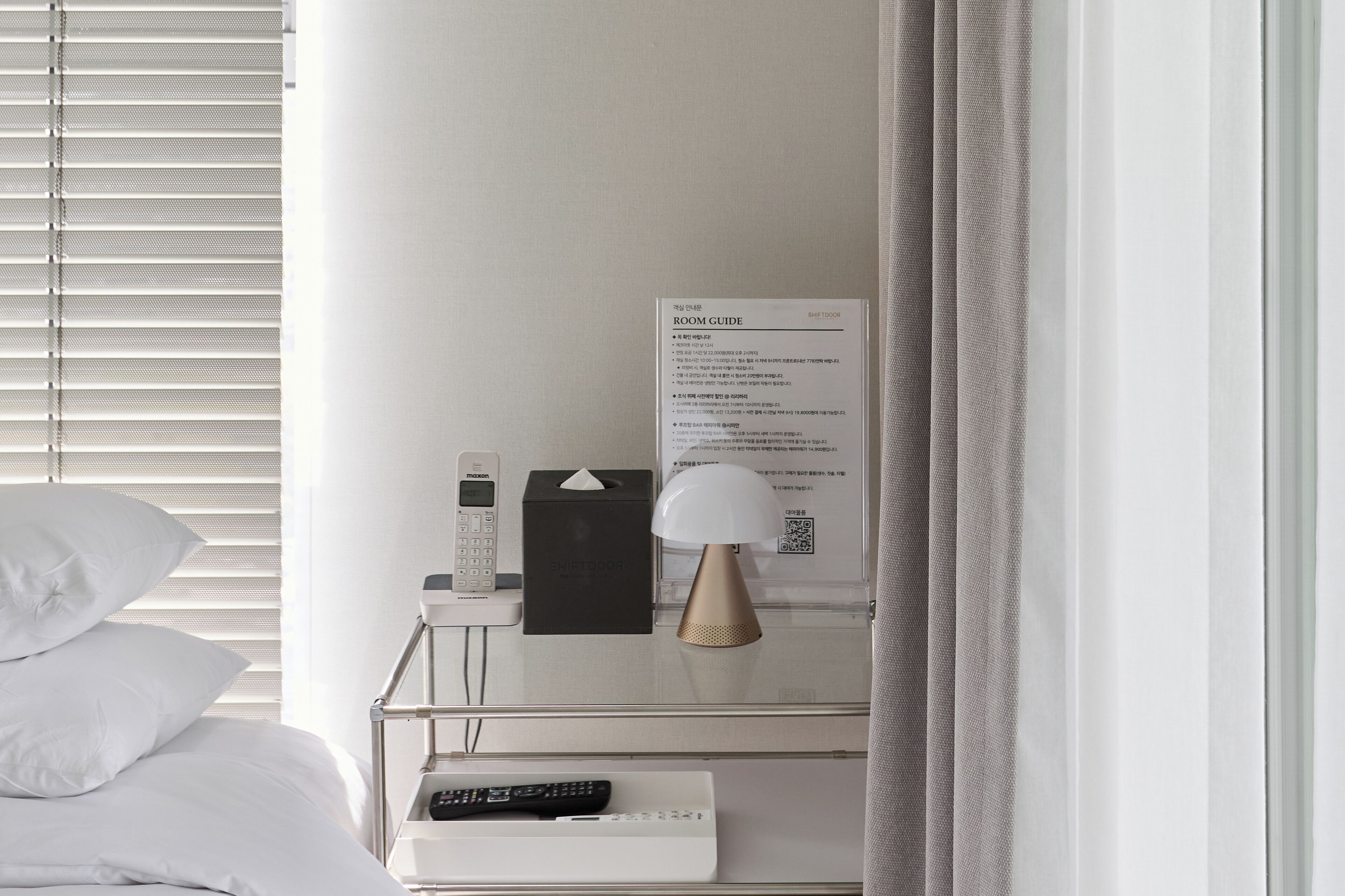 A hotel room with a bed, a bedside table with a lamp, a remote control, a tissue box, and a room guide on the wall.