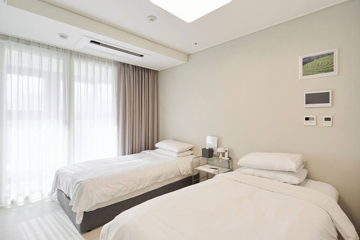 A hotel room with two beds, a bedside table with a lamp, a wall-mounted TV, and a window with curtains.