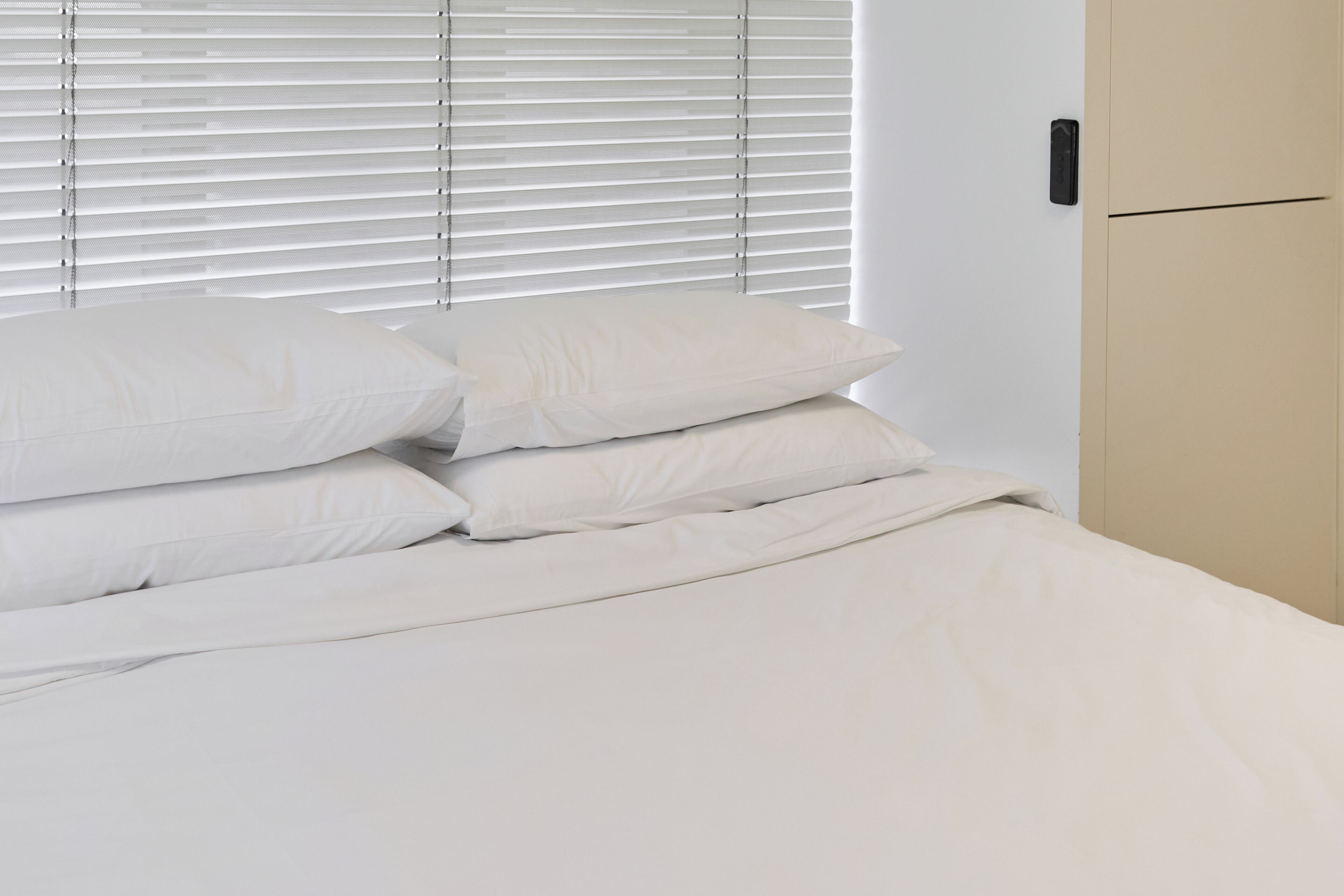 A neatly made bed with white linens and pillows, a closed door, and a window with blinds.