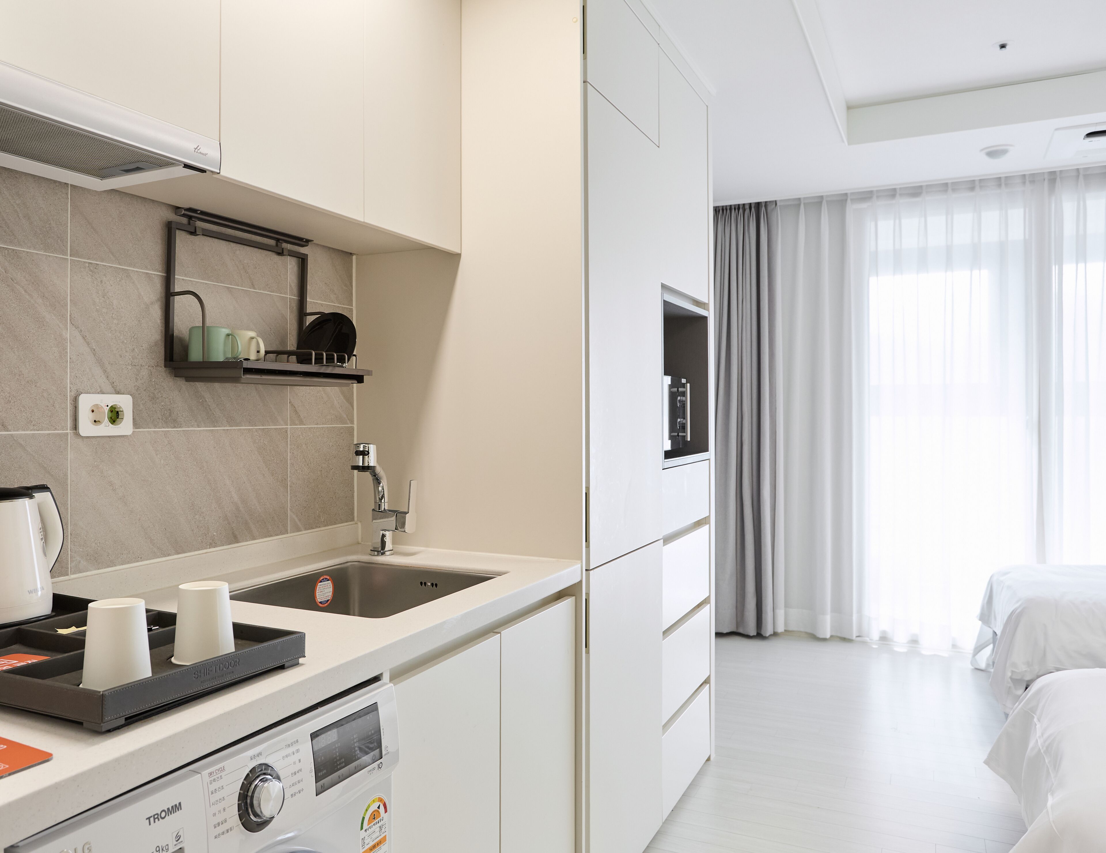 A modern hotel room with a kitchenette, a large window with curtains, and a bed.