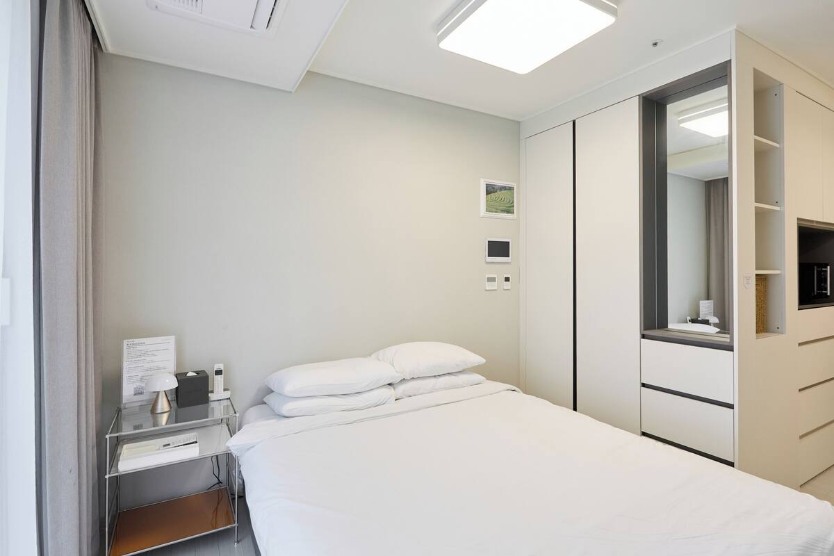 A modern hotel room with a neatly made bed, a bedside table with a lamp, a small shelf, and a wardrobe with a mirror.
