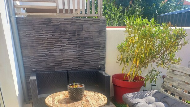 A patio area with a round wooden table, two wicker chairs, a red potted plant, and a grey cushioned bench. - Lovely 3-bedroom family home for rent Ideal location (La Rochelle)
