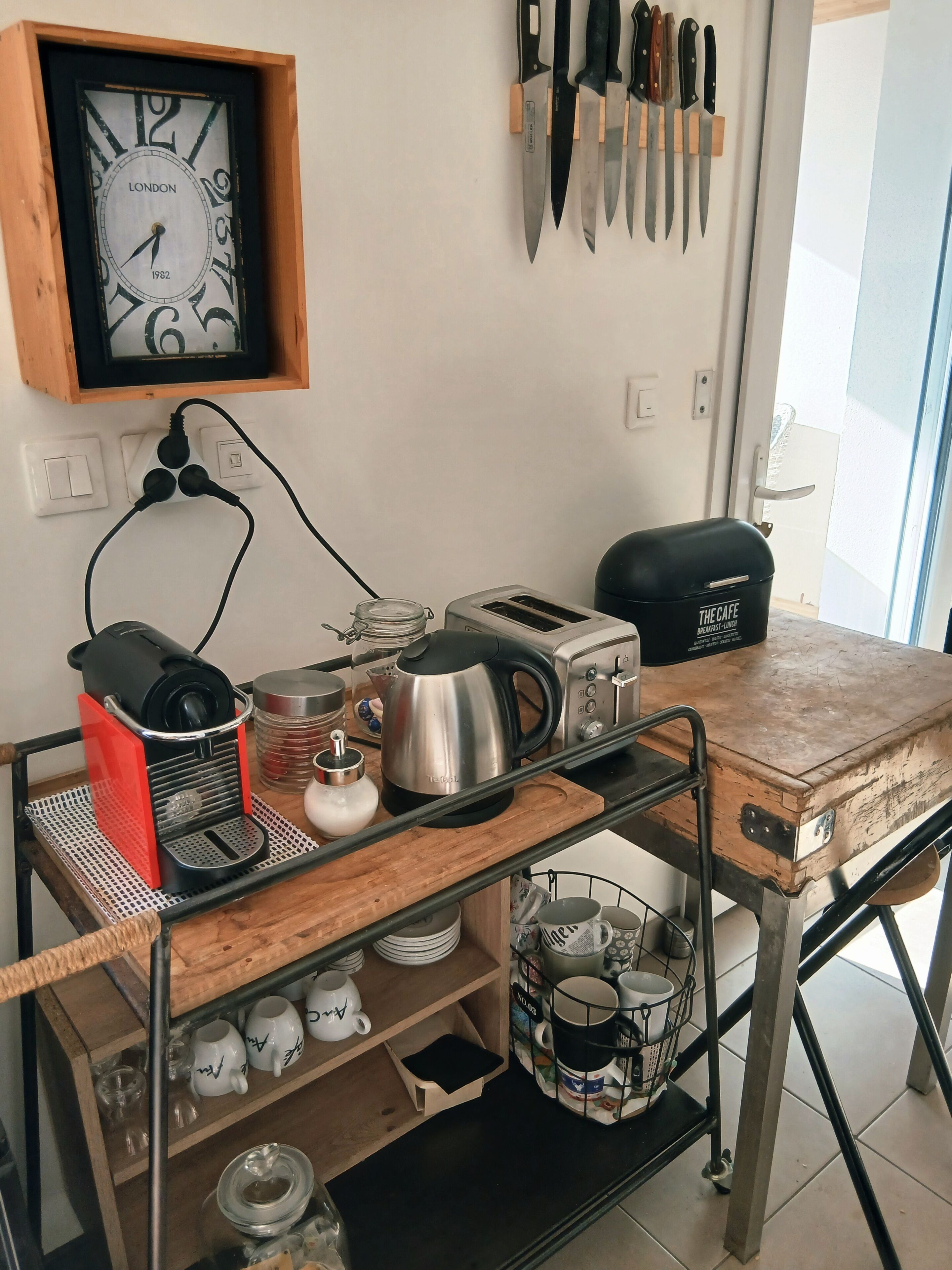A kitchen area with a coffee maker, toaster, kettle, and various mugs on a wooden cart.