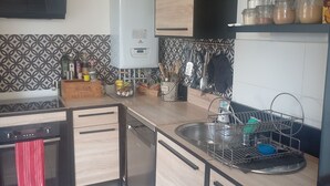 A modern kitchen with a built-in dishwasher, a gas stove, and a sink with a metal rack. There are jars on the countertop and a potted plant on the shelf. - Lovely 3-bedroom family home for rent Ideal location (La Rochelle)
