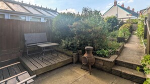 Un patio con una mesa y sillas de madera, una jardinera de ladrillo con una fuente de agua antigua, y un jardín con diversas plantas.