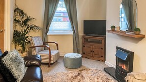 Una sala de estar acogedora con un sofá de cuero, un sillón a rayas, un tapete mullido, un reposapiés pequeño y redondo, un televisor sobre un mueble de madera, una chimenea y un espejo grande en la pared.