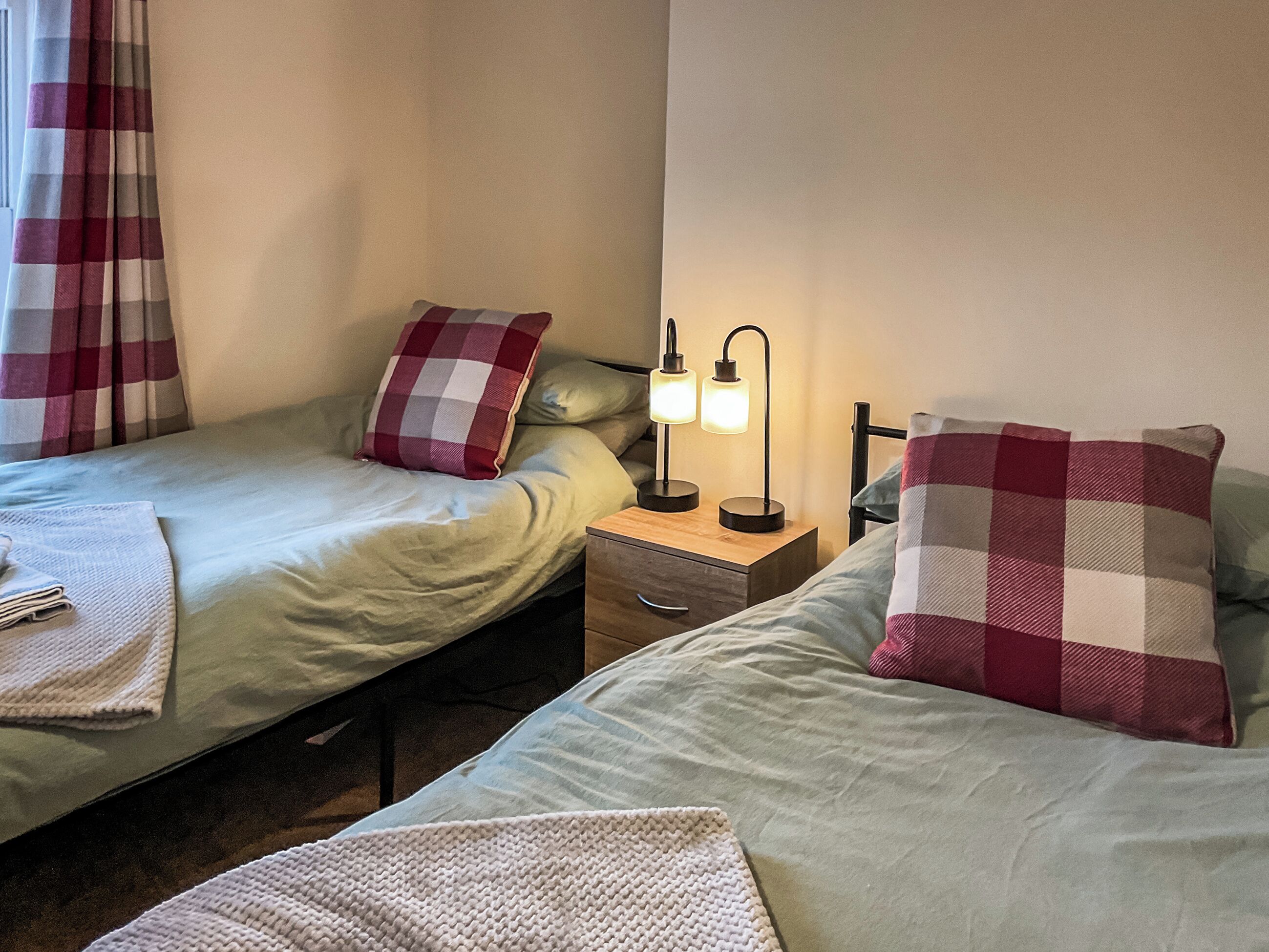 A twin room with two beds, each with a checkered pillow, a bedside table with a lamp, and a folded towel on the floor.