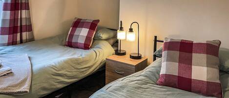 A twin room with two beds, each with a checkered pillow, a bedside table with a lamp, and a folded towel on the floor.