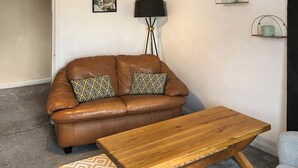 Una sala de estar con un sofá de cuero marrón, dos cojines estampados, una mesa de centro de madera, una lámpara de pie y un estante pequeño montado en la pared con objetos decorativos.