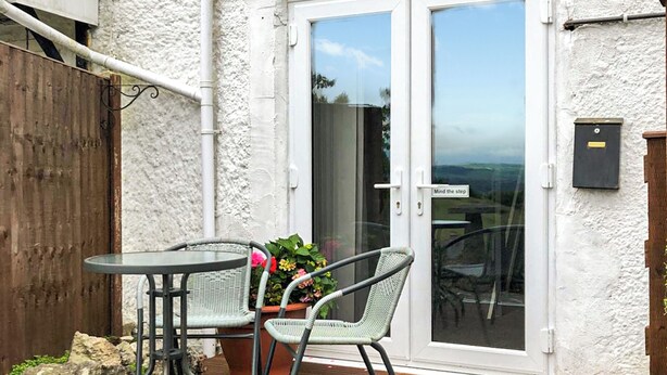 Un patio exterior con una mesa y dos sillas, una terraza de madera, una pared blanca con textura y una puerta corredera de vidrio con un letrero que dice 'Mind the step'.
