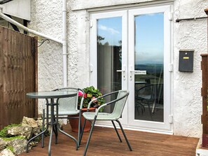 Eine Terrasse im Freien mit Tisch und zwei Stühlen, ein Holzdeck, eine weiß gestreichte Wand und eine Glastür mit einem Warnschild 'Achtung trittstufe'.
