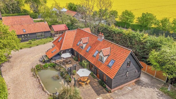 A house with a sloped roof, a patio with outdoor furniture, and a pond. - 7 bedroom accommodation in Ilketshall St Margaret near Bungay (Ilketshall St Margaret near Bungay)