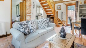 A living room with a sofa, a wooden table, a staircase, and a kitchen in the background. - 7 bedroom accommodation in Ilketshall St Margaret near Bungay (Ilketshall St Margaret near Bungay)