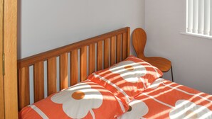 A bedroom with a wooden headboard, a single bed with an orange and white floral bedspread, and a window with vertical blinds.