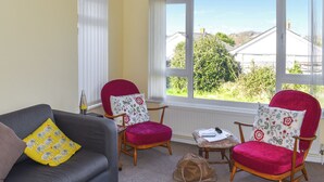A room with a grey sofa, two red armchairs, a small table, and a window with a view of houses and greenery.