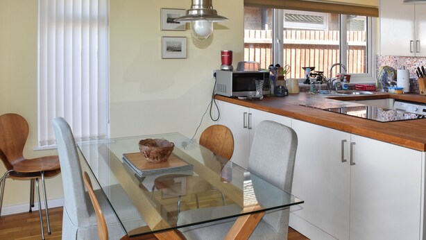 A modern kitchen with a glass dining table, wooden chairs, and white cabinets.
