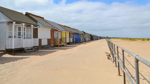 A row of beach huts with a sandy path leading to the beach, a metal railing along the path, and a clear sky above.