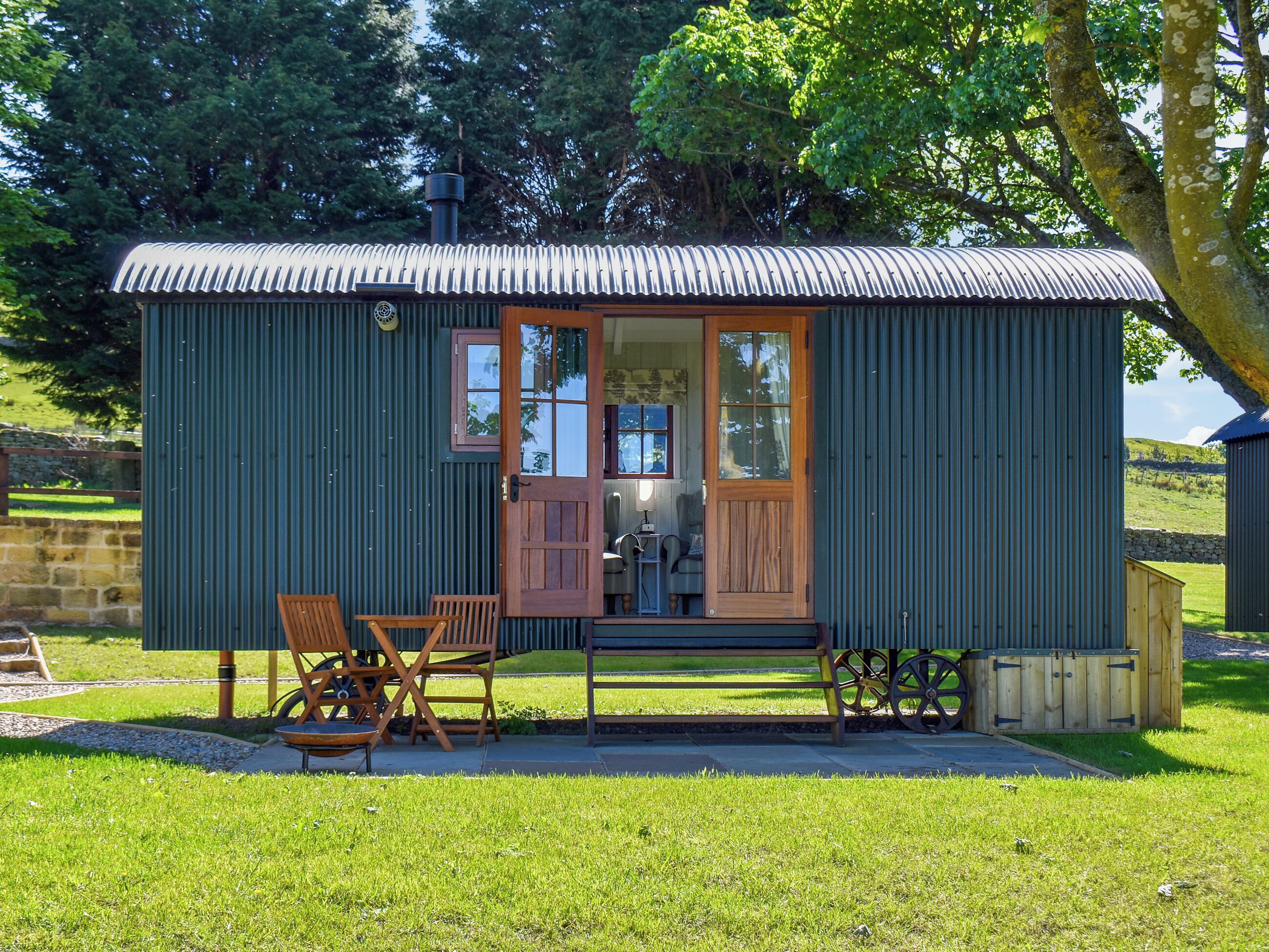 A small, single-story building with a corrugated metal roof and a wooden door. It features a small porch with a bench and a table, surrounded by a grassy area.