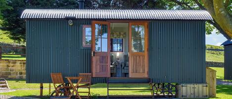 A small, single-story building with a corrugated metal roof and a wooden door. It features a small porch with a bench and a table, surrounded by a grassy area.