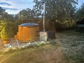 A wooden hot tub with a metal chimney, surrounded by potted plants and a stone pathway.