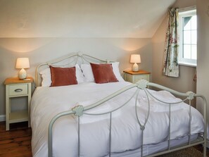 Una cama bien tendida con ropa blanca y dos almohadas marrones, flanqueada por dos mesitas de noche con lámparas, en una habitación con ventana y cortinas florales.