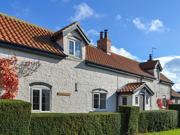 A white brick cottage with a red-tiled roof, a small attached garage, and a well-maintained hedge.