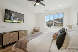A bedroom with a bed, a television mounted on the wall, a ceiling fan, and a window with a view of houses. - Heated Pool Oasis by the Santa Rosa Mountains (La Quinta)