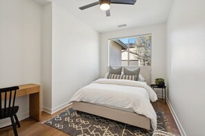 A bedroom with a bed, a desk, a chair, a ceiling fan, and a window with a view of the outside.