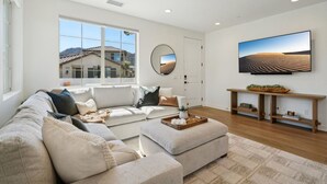 A modern living room with a sectional sofa, a coffee table, a flat-screen TV, and a large window offering a view of a house and mountains. - Heated Pool Oasis by the Santa Rosa Mountains (La Quinta)