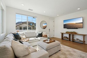 A modern living room with a sectional sofa, a coffee table, a flat-screen TV, and a large window offering a view of a house and mountains. - Heated Pool Oasis by the Santa Rosa Mountains (La Quinta)