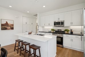 A modern kitchen with white cabinetry, a central island with stools, stainless steel appliances, and a desert landscape painting on the wall.
